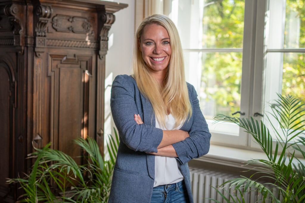 Das Bild zeigt Stina Ehrecke - Chief Executive Officer – stehend vor einem hellen Fenster am rechten Bildrand, dunklen Holzelementen am linken und Palmwedeln, die von unten ins Bild ragen. Sie schaut lächelnd in die Kamera.