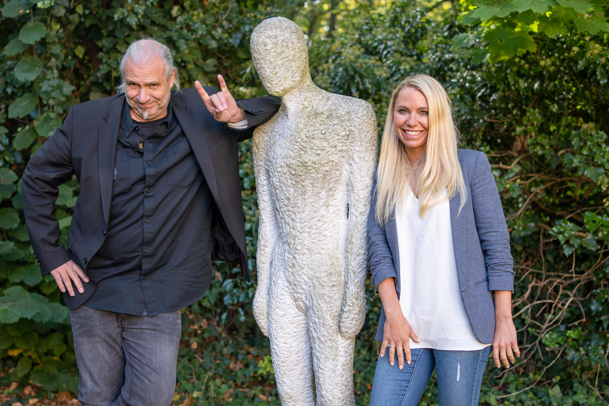 Das Bild zeigt lächelnd und mit Blick zur zeigt Kamera gerichtet links Dipl.-Ing. Uwe Lambrecht rechts Stina Ehrecke. Zwischen Ihnen steht eine Steinskulptur in menschlicher Gestalt. Der Hintergrund besteht komplett aus dem Grün von Pflanzen und Büschen.
